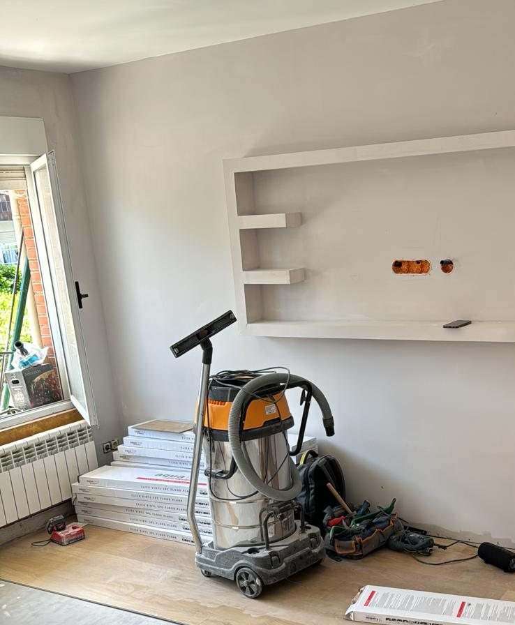 Estancia con paredes pintadas en gris claro, estantería de obra, cajas de suelo vinílico y aspirador de obra