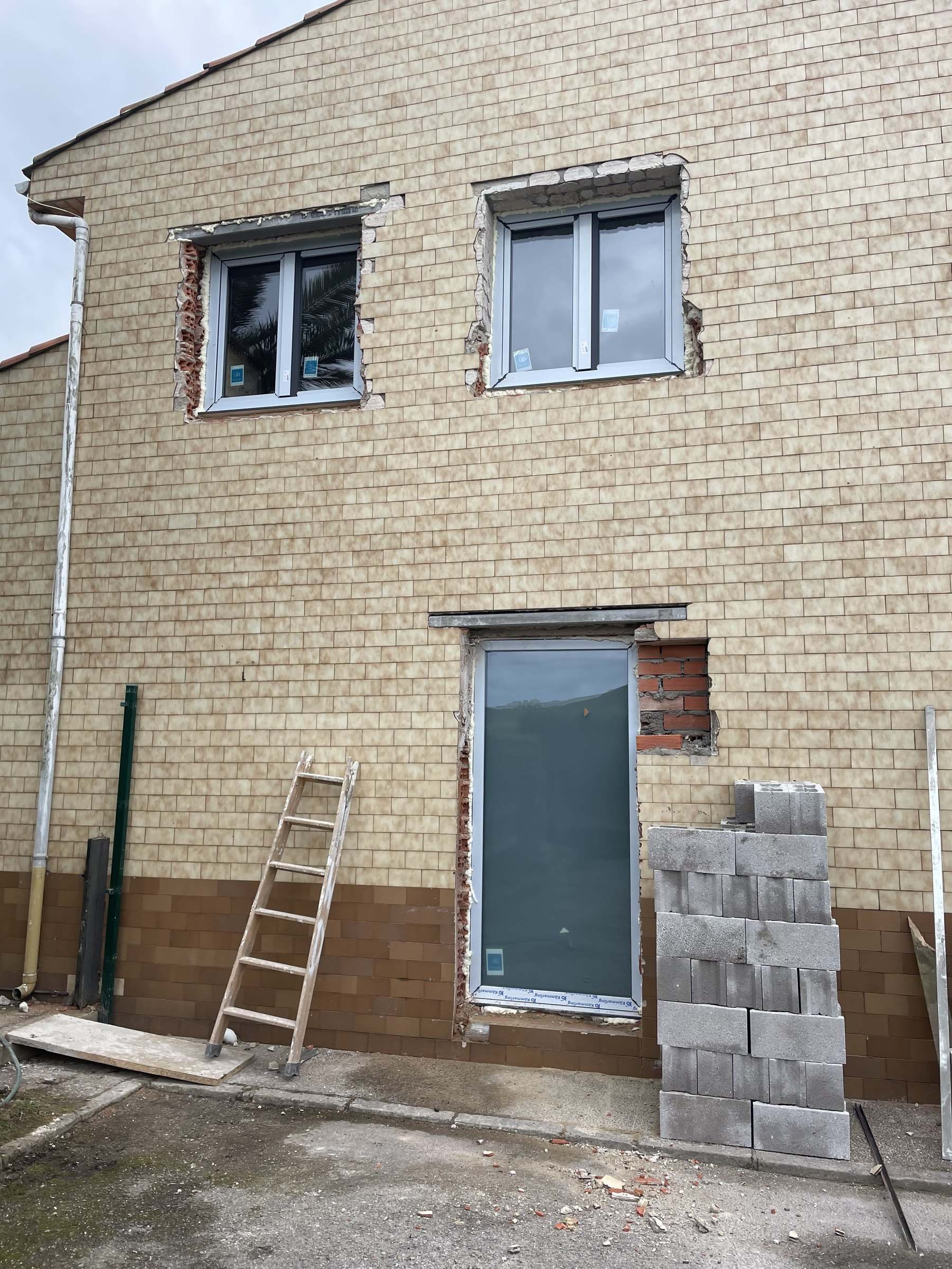 Fachada lateral en obra: puerta nueva con el hueco sin rematar, ladrillo a la vista y bloques de hormigón apilados
