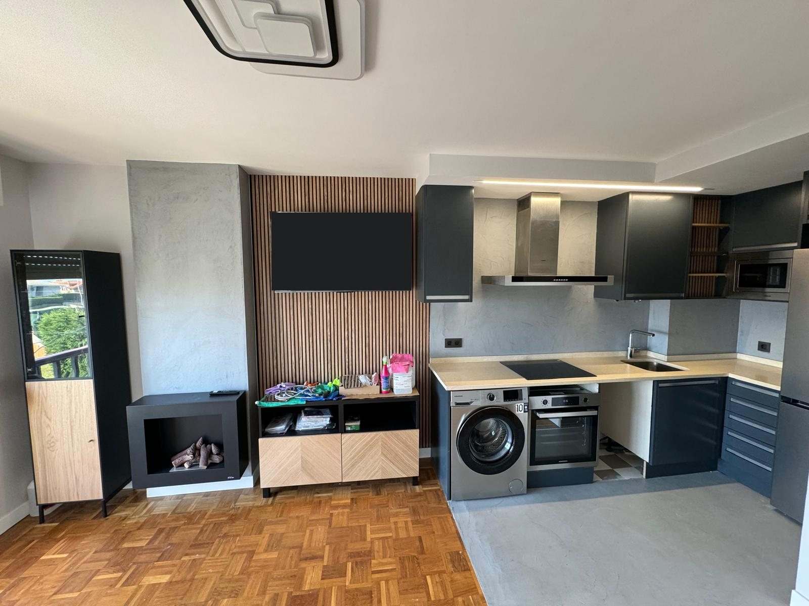 Salón-cocina terminado: cocina en L antracita, panel de listones de madera con televisión, pilar gris y parquet