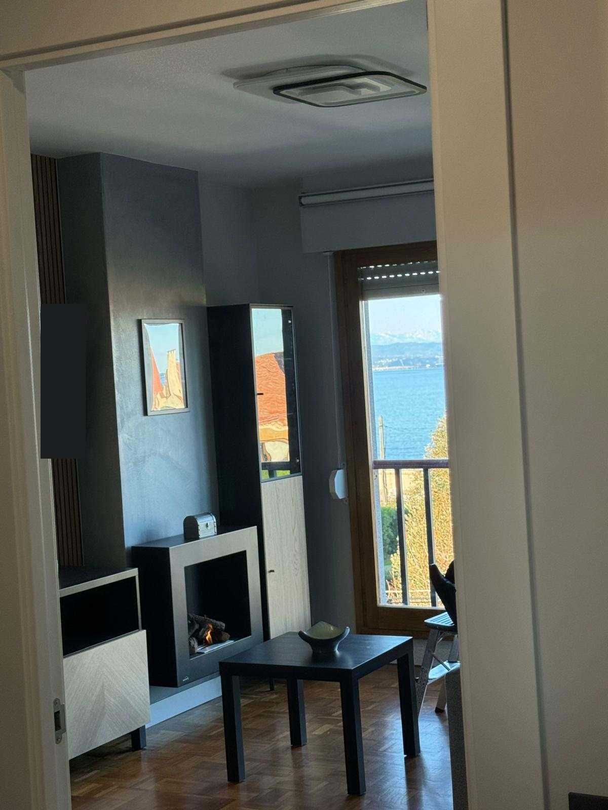 Rincón del salón con chimenea decorativa, mesa baja y puerta de terraza con vistas al mar