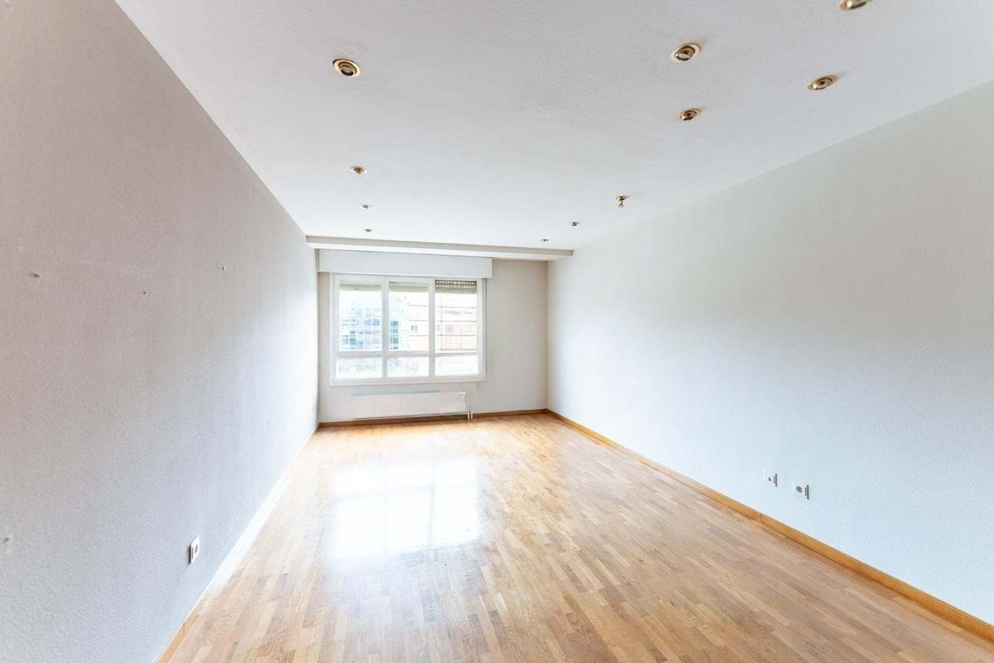 Salón vacío con suelo de parquet, paredes claras y ventanal al fondo